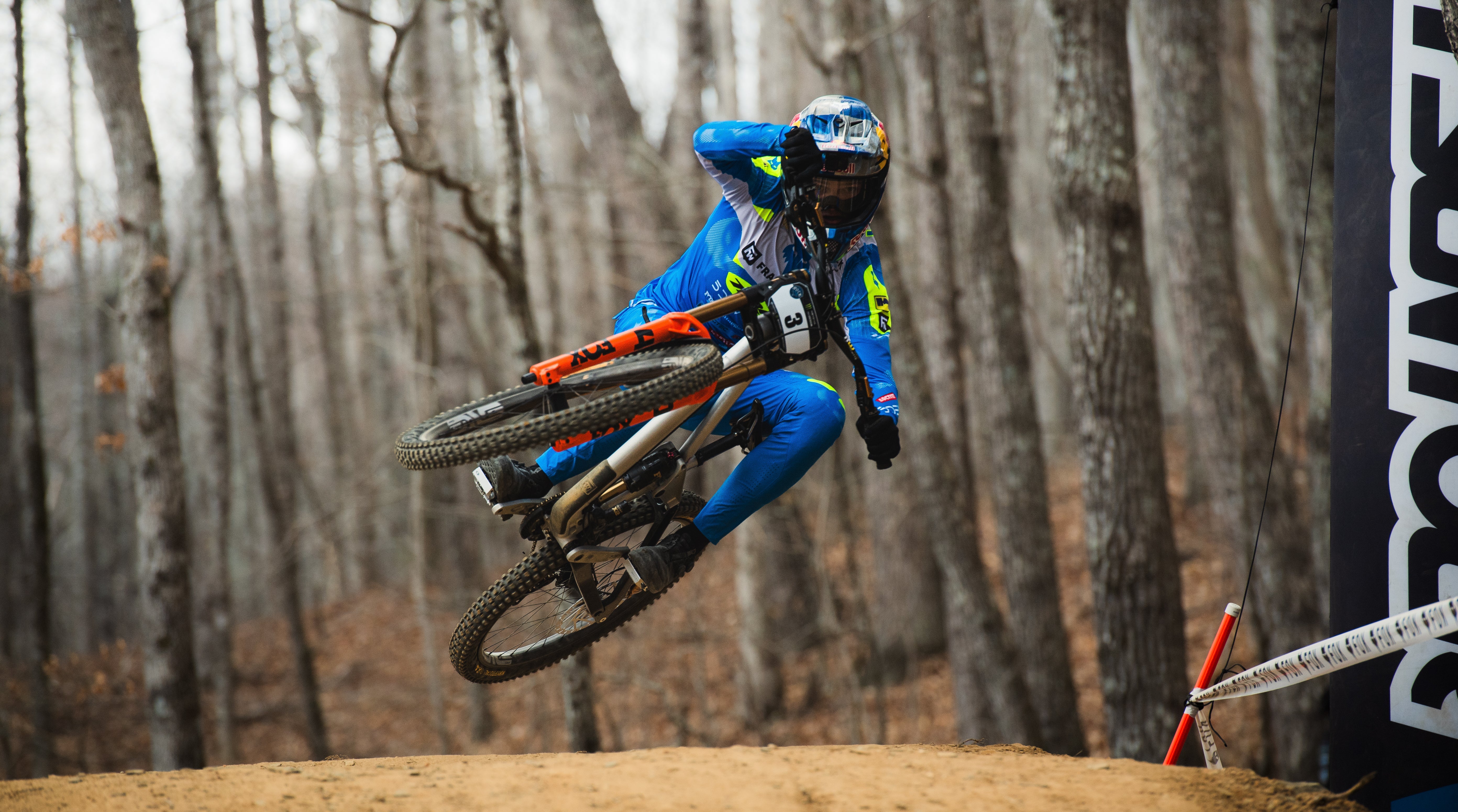 Biker in blue jacket and pants performing a jump on a dirt bike in a forest setting.