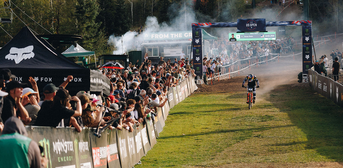 A mountain biker rides through a dusty finish line at an outdoor race, with a large crowd of spectators lining the track behind barriers. Fox branding is visible on tents and signage, and smoke fills the background near the arch.