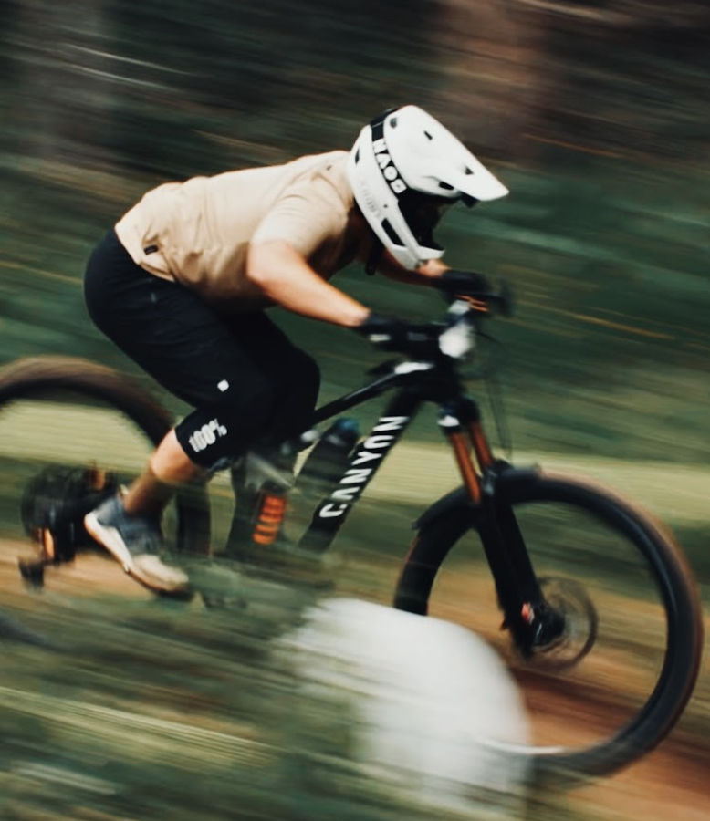 A mountain biker in a beige shirt, black shorts, and a white full-face helmet rides a black Canyon mountain bike on a blurred forest path.