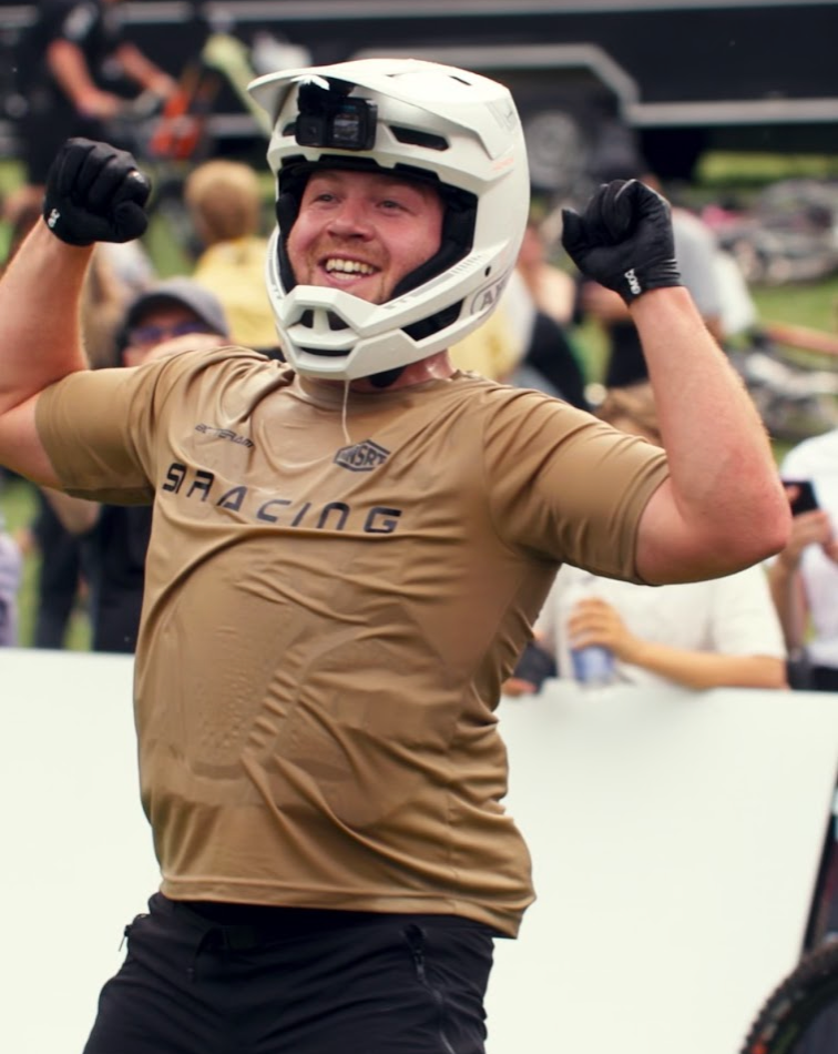 A smiling man wearing a white full-face helmet with a mounted camera and a tan short-sleeve jersey with 'SRACING' across the chest, raising both arms in celebration. The background shows a blurred outdoor crowd and event setting.