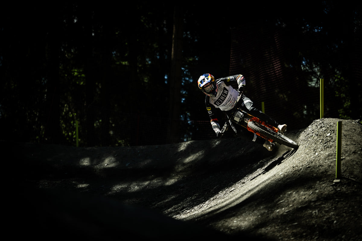 A motocross rider in white and black gear leans sharply into a turn on an orange and black bike over a dirt track, with dark trees in the background.