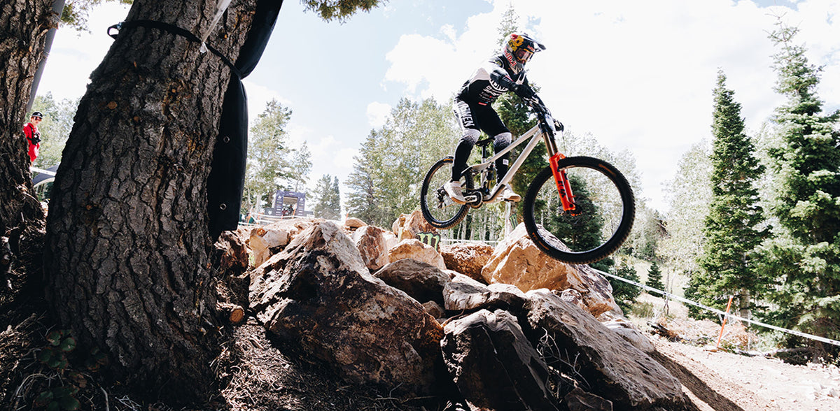 A mountain biker in full gear jumps a white and red bike over large rocks in a forest setting, with a tree trunk prominent on the left side of the frame.