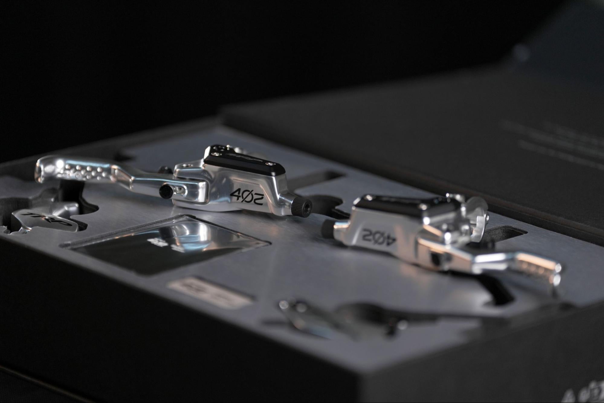 Close-up of two metallic bicycle pedal components resting in a custom-cut black presentation box, with the number 402 engraved on each piece.