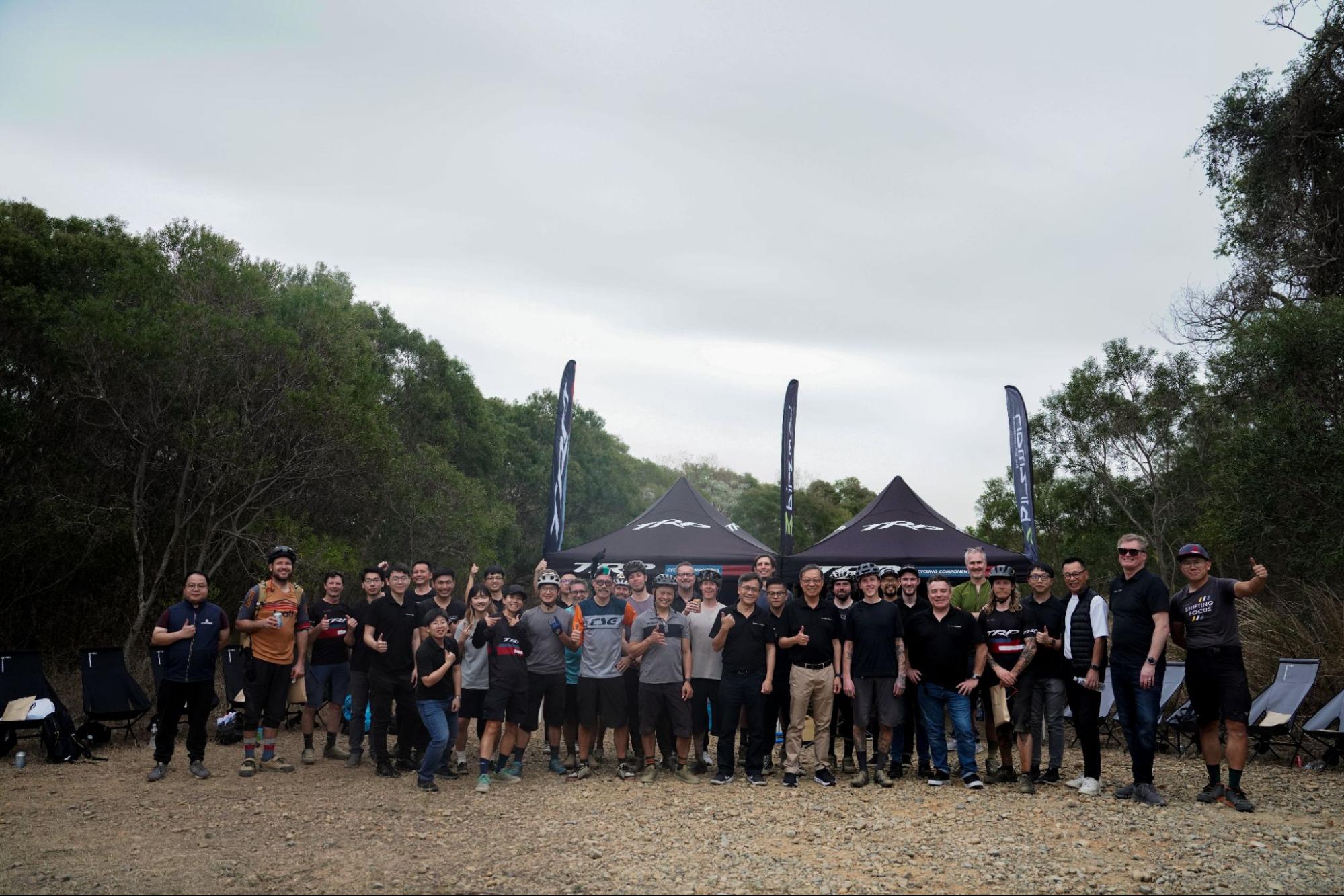 A large group of people posing together outdoors in front of two black event tents, with trees and an overcast sky in the background.