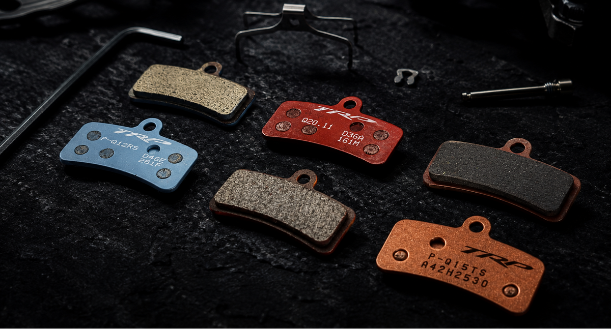 Five brake pads in different colors (blue, red, black, brown, copper) arranged on a dark textured surface with a hex key and mounting tool in the background.