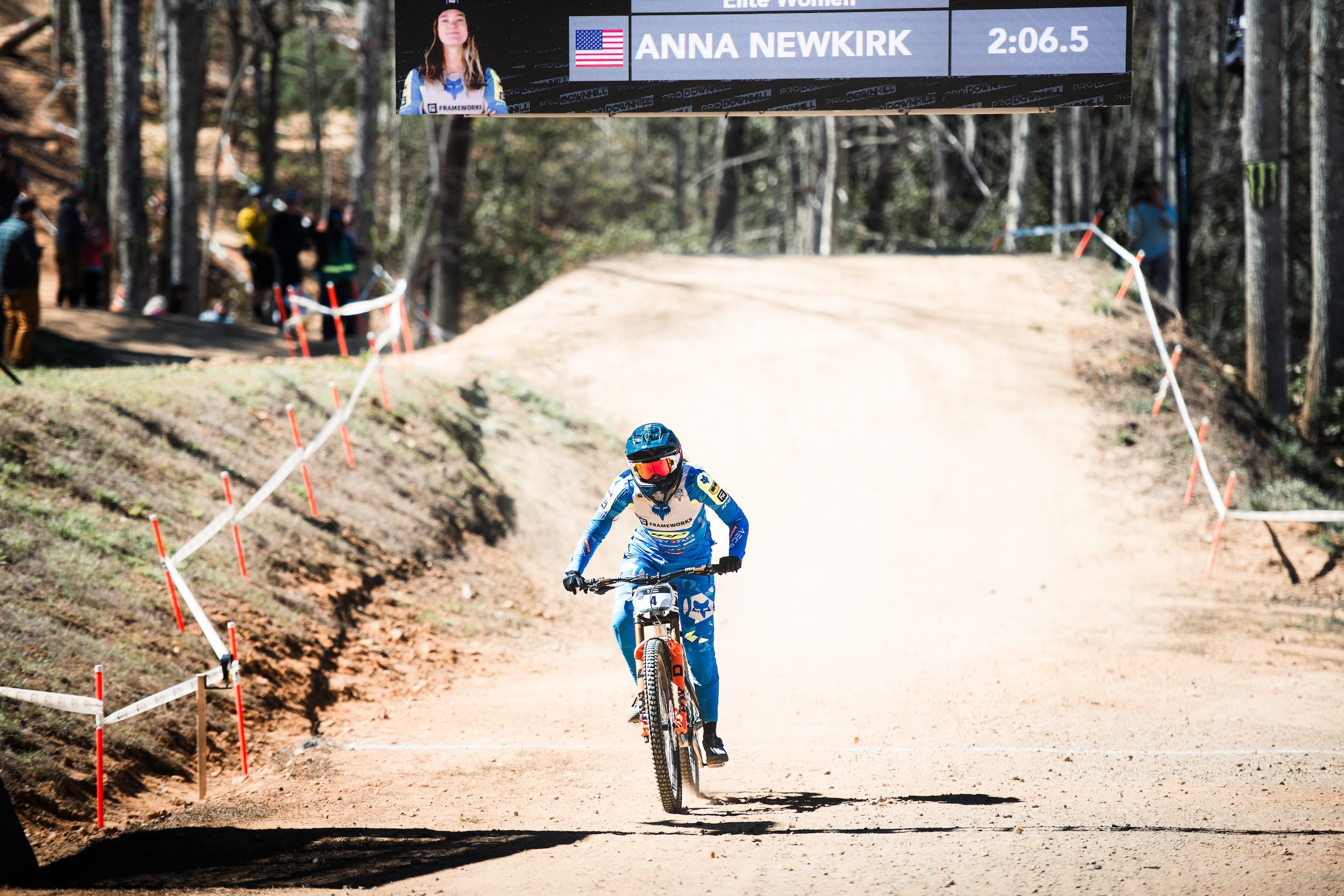 Biker in blue jersey and helmet riding a dirt track with 'Anna Newkirk' sign above.