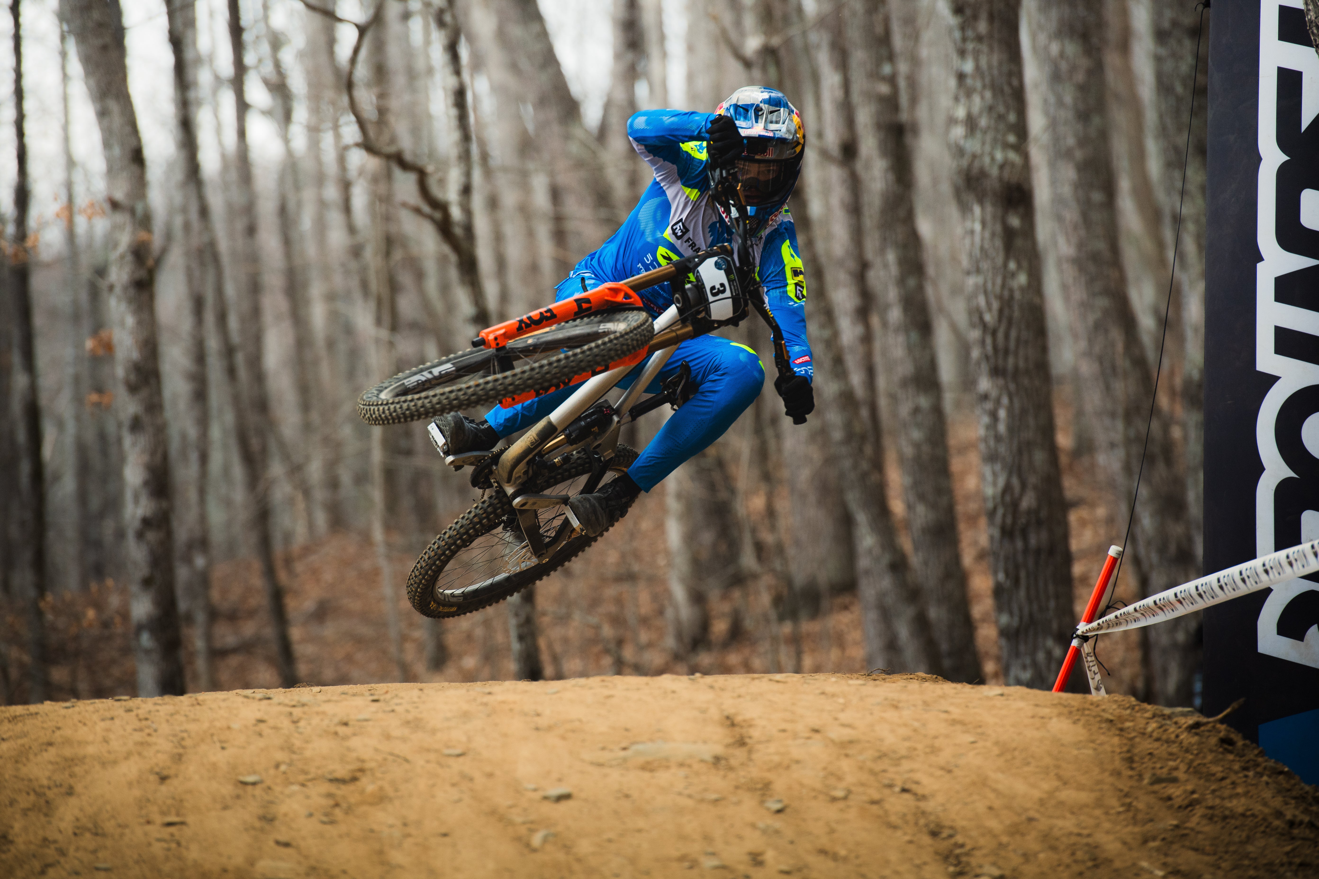 Biker in blue jacket and pants performing a jump on a dirt bike in a forest setting.