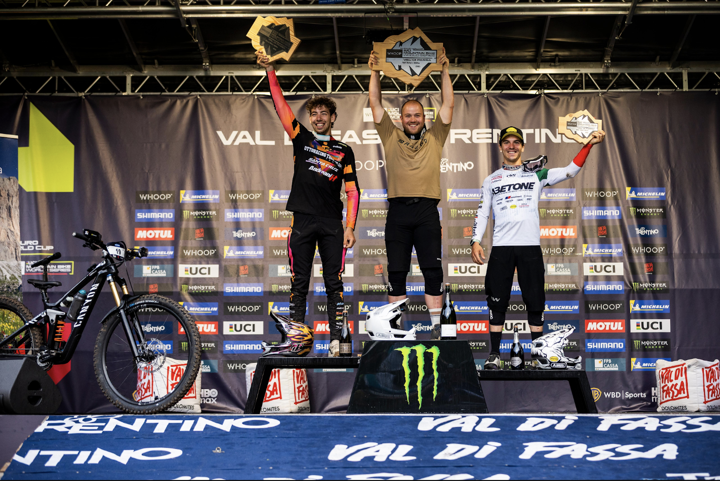 Three mountain bike riders stand on a podium at the Val di Fassa International Raceway, holding gold trophies. The rider on the left wears a black and orange jersey with a black and orange bike, the center rider wears a tan jersey with a tan bike, and the rider on the right wears a white and green Visotino jersey with a white and green bike.