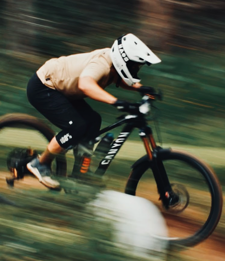 A mountain biker in a beige shirt, black shorts, and a white full-face helmet rides a black Canyon mountain bike on a blurred forest path.