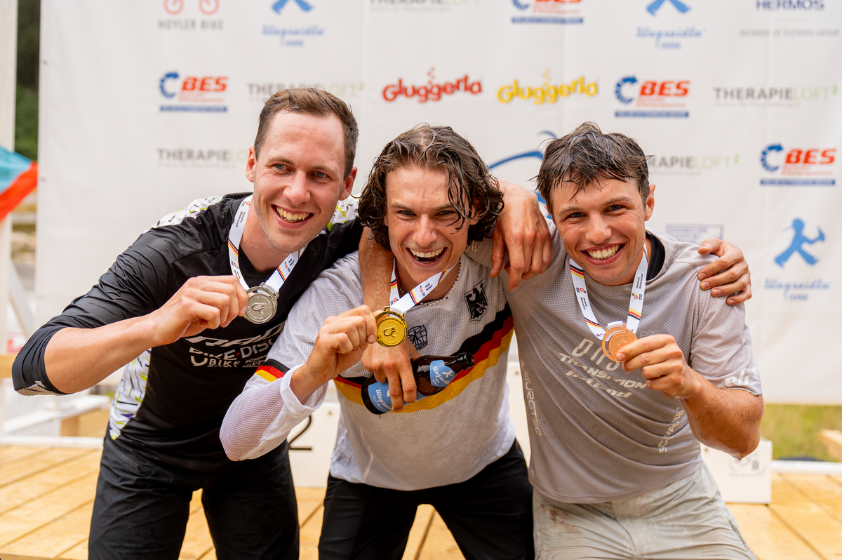 Three male athletes celebrating on a podium with medals, wearing sports jerseys and posing in front of a branded sponsor backdrop.
