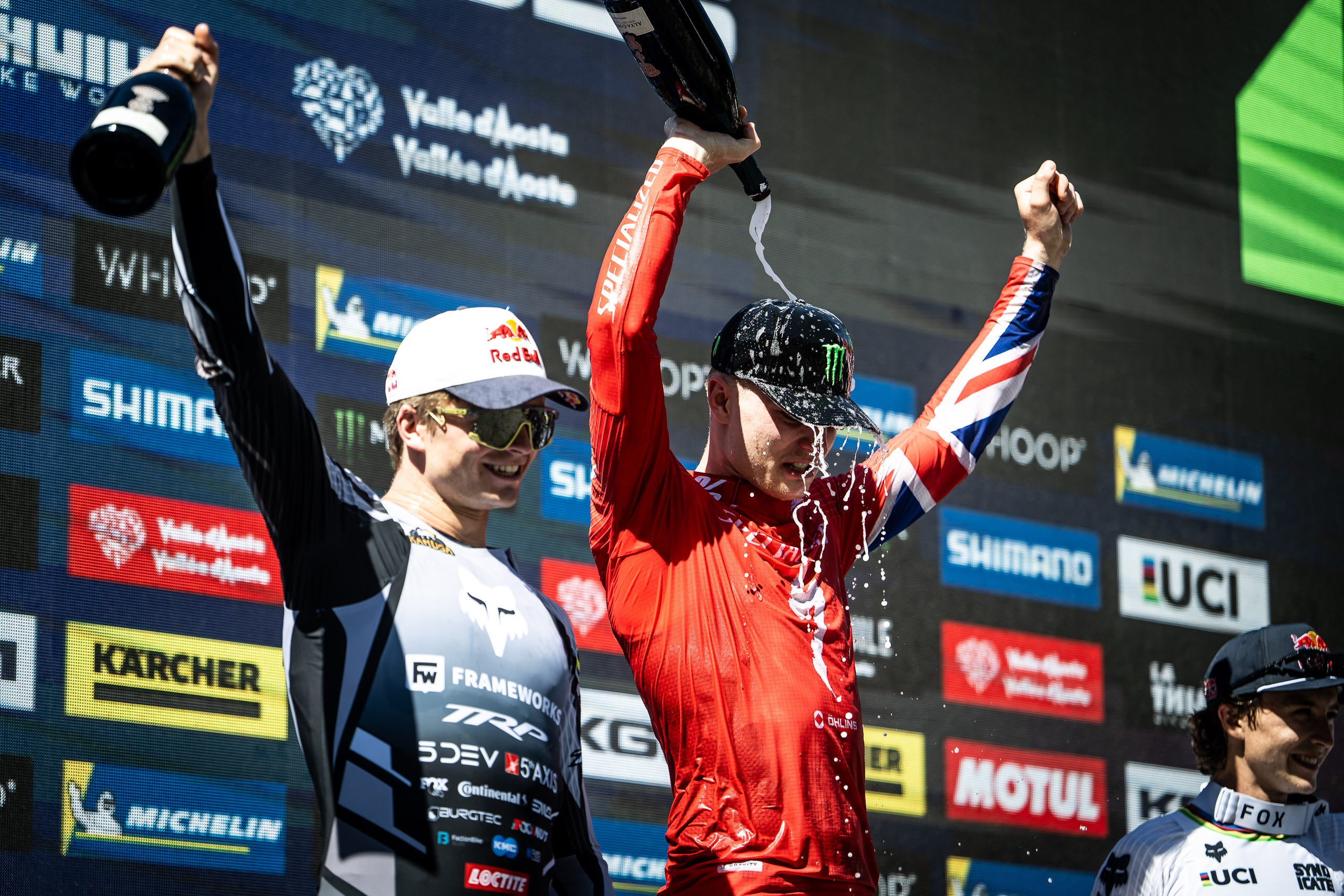 Two athletes on a podium celebrating with champagne bottles, one in a white and black Framewinds jersey and one in a red long-sleeve top with Union Jack details, spraying liquid on each other against a sponsor backdrop.