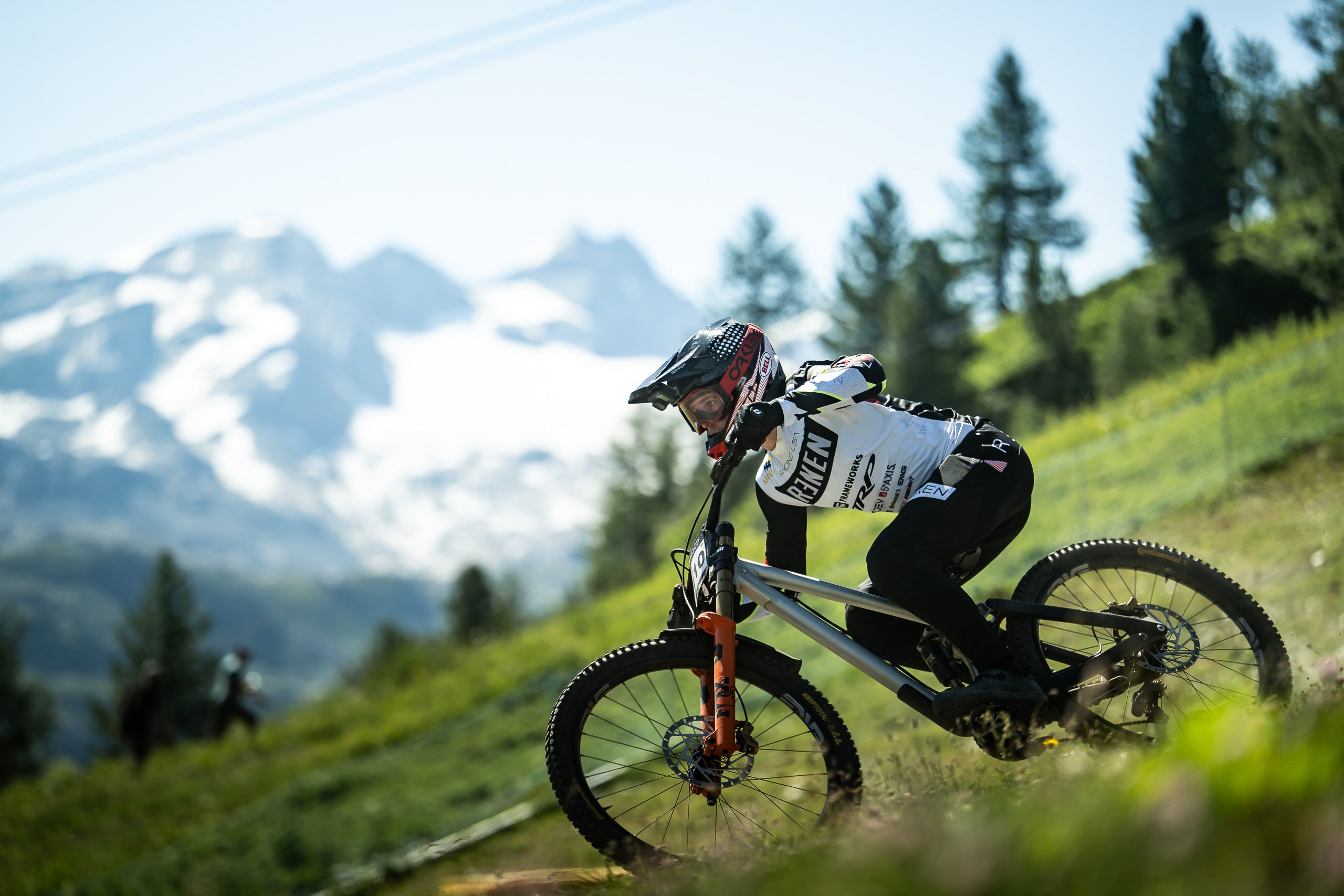 A mountain biker in a white and black jersey leans sharply on a full-suspension downhill bike on a grassy trail, with snow-capped mountains and pine trees in the background.