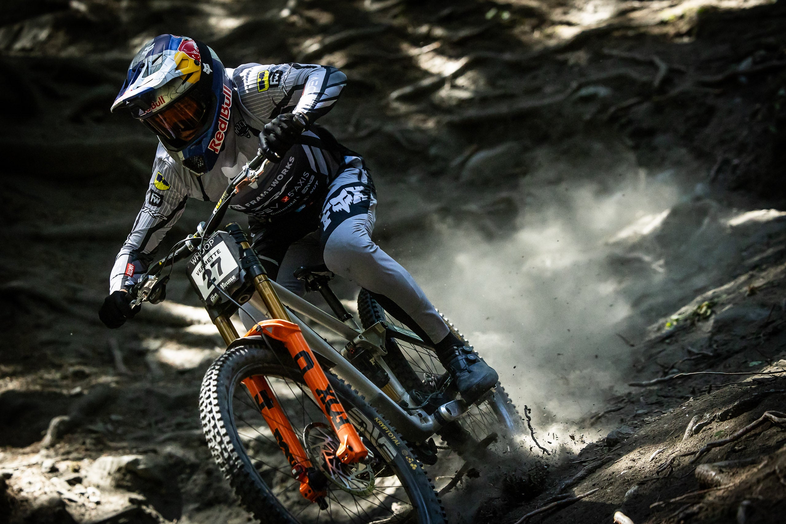 A mountain biker in a white, black, and blue racing suit leans sharply into a turn on a dirt trail, kicking up dust. The bike has an orange front fork and a visible Fox suspension brand.