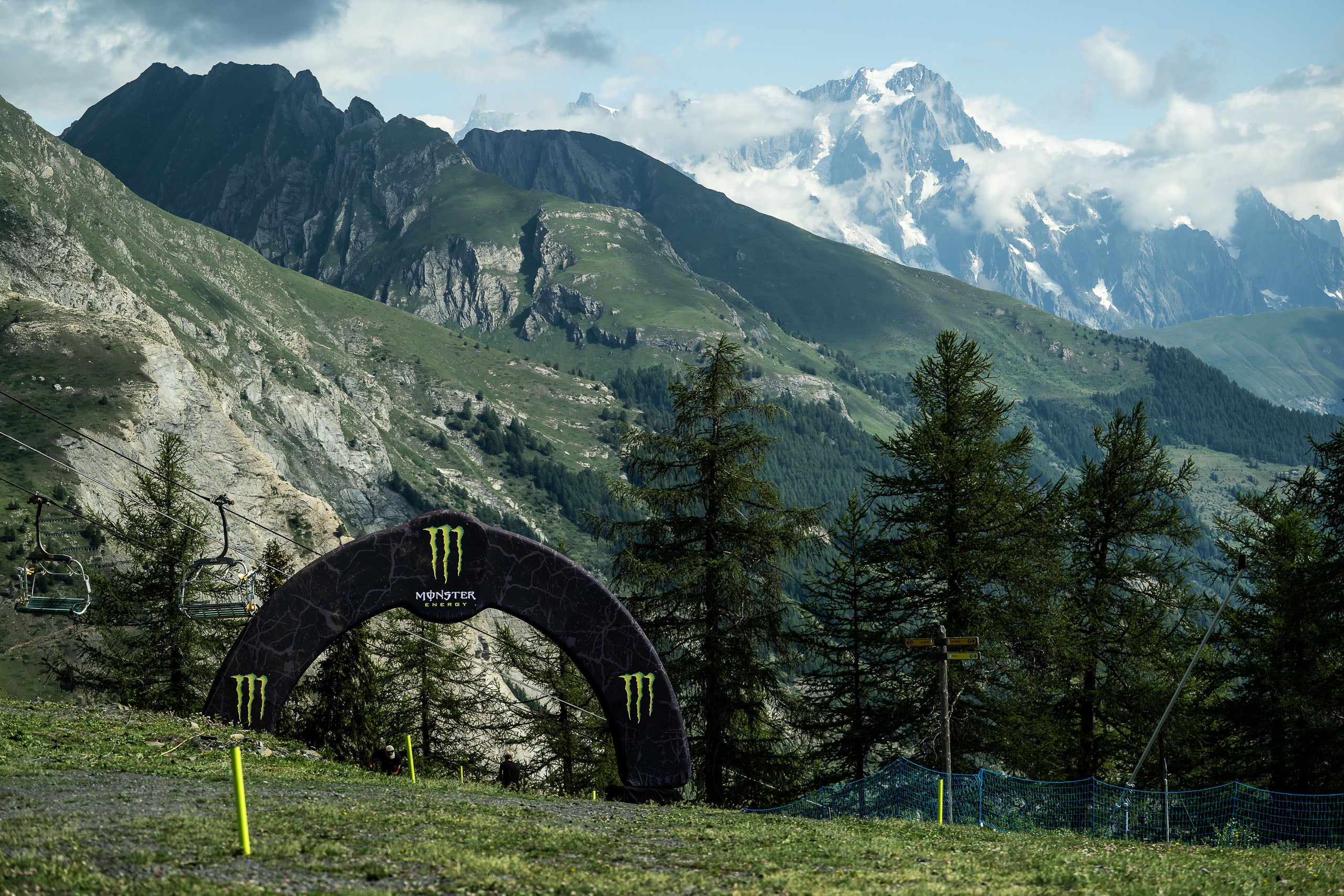 A black Monster Energy archway with bright green claw logos stands in a grassy mountain clearing, with evergreen trees and rugged alpine peaks in the background under a cloudy sky.