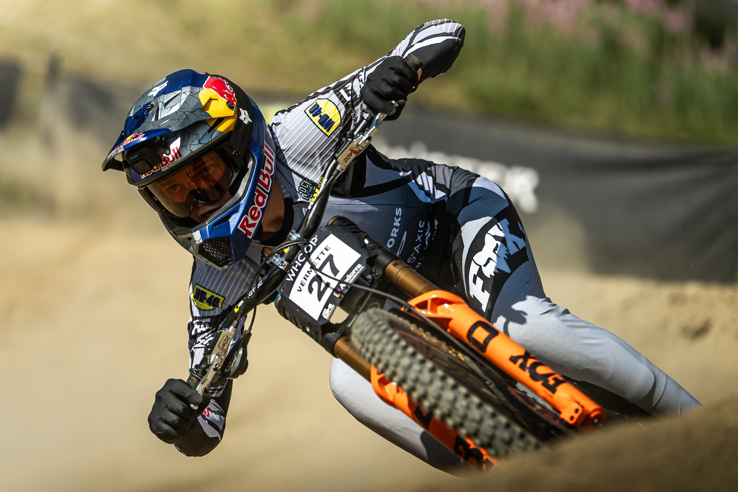 A motocross racer leans sharply into a turn on an orange and black Fox dirt bike, wearing a blue, white, and black Red Bull helmet and matching sponsor apparel on a dirt track.