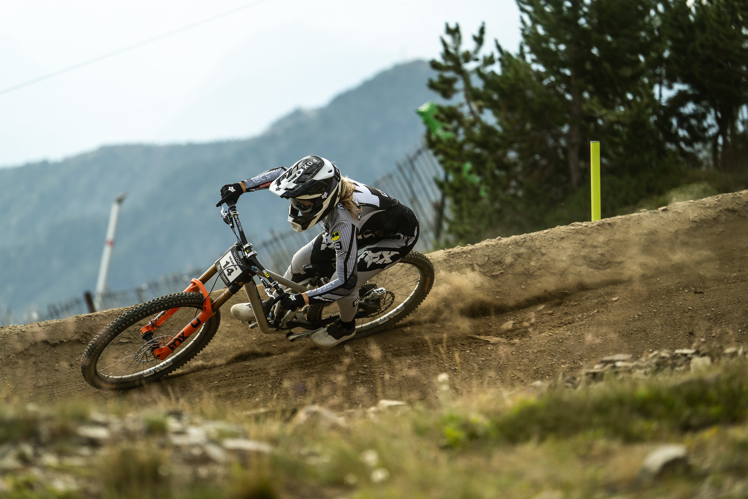 A mountain biker in full racing gear leans sharply into a turn on a dirt track, with trees and mountains blurred in the background.