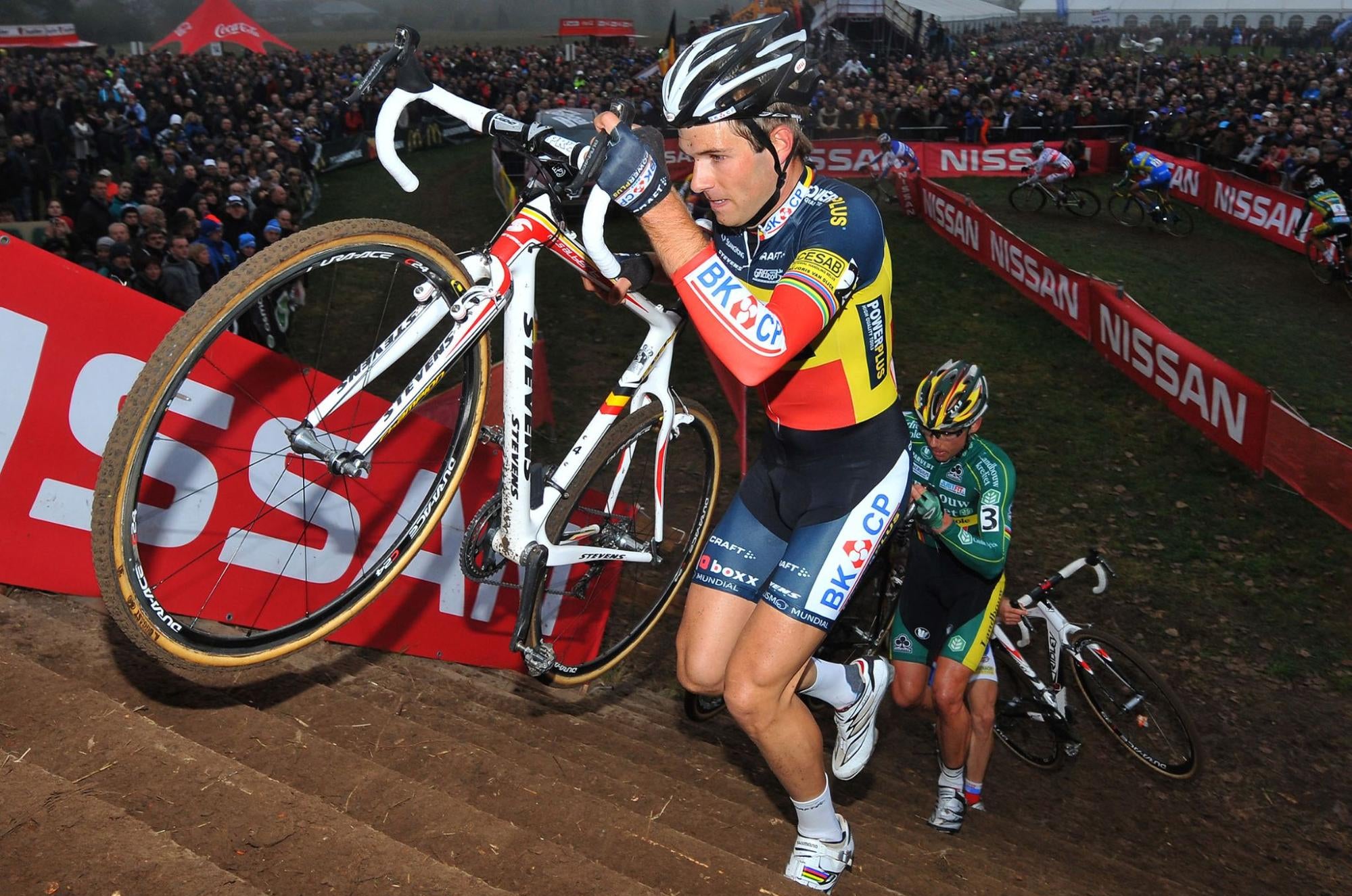 A cyclist in a blue, yellow, and red BIC CP jersey struggles up muddy stairs while carrying a white road bicycle. Another cyclist is behind him.