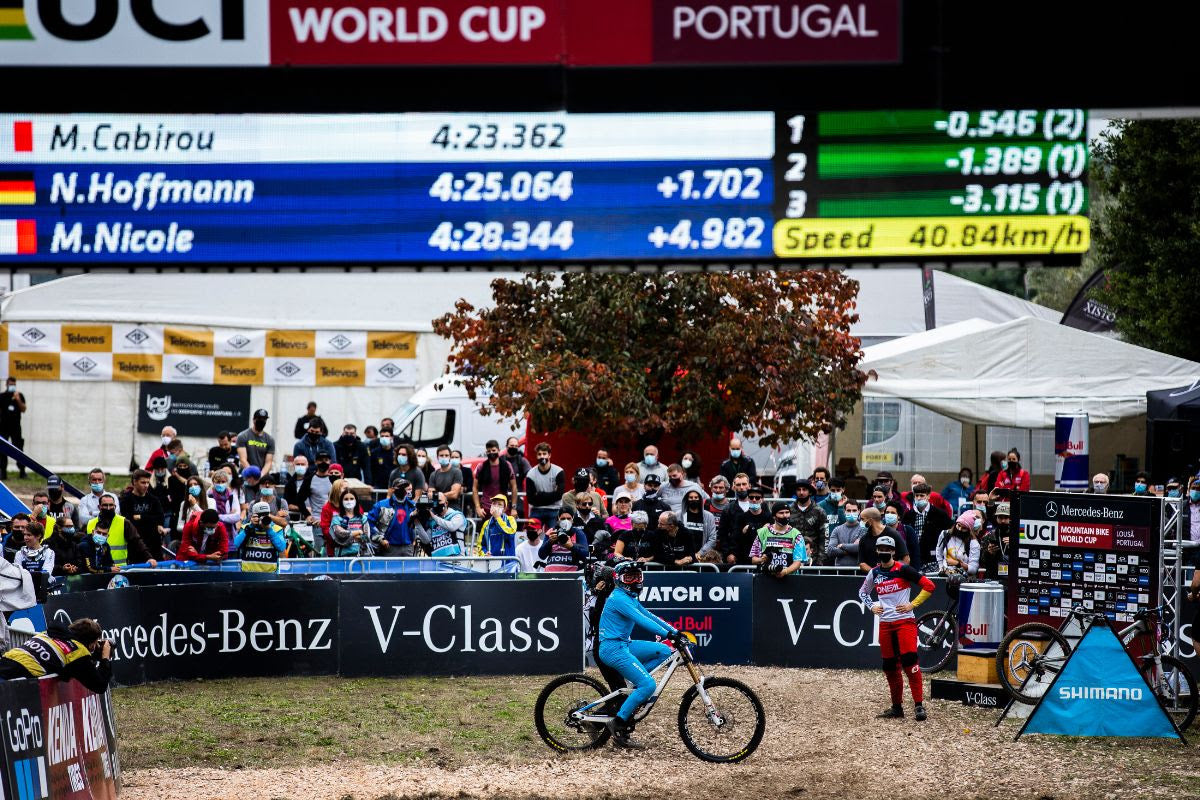 A cyclist rides a mountain bike in front of a large race scoreboard at a V-Class World Cup event in Portugal. The scene includes a crowd of spectators, race barriers, and visible sponsor logos such as Mercedes-Benz, Shimano, and Vacant.