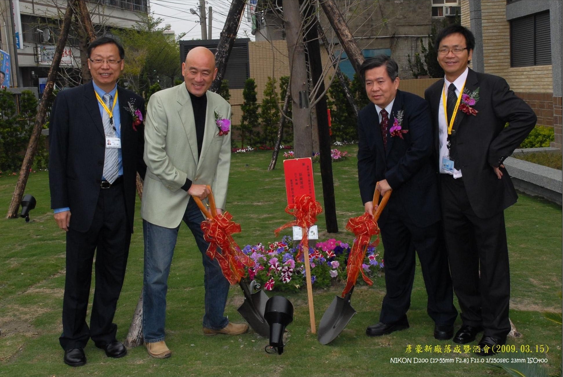 Four men standing outdoors on grass in front of trees and buildings, holding shovels with red ribbons near a small plaque and flower arrangement. Two men wear dark suits and two wear light-colored suits; one man wears a light green blazer.
