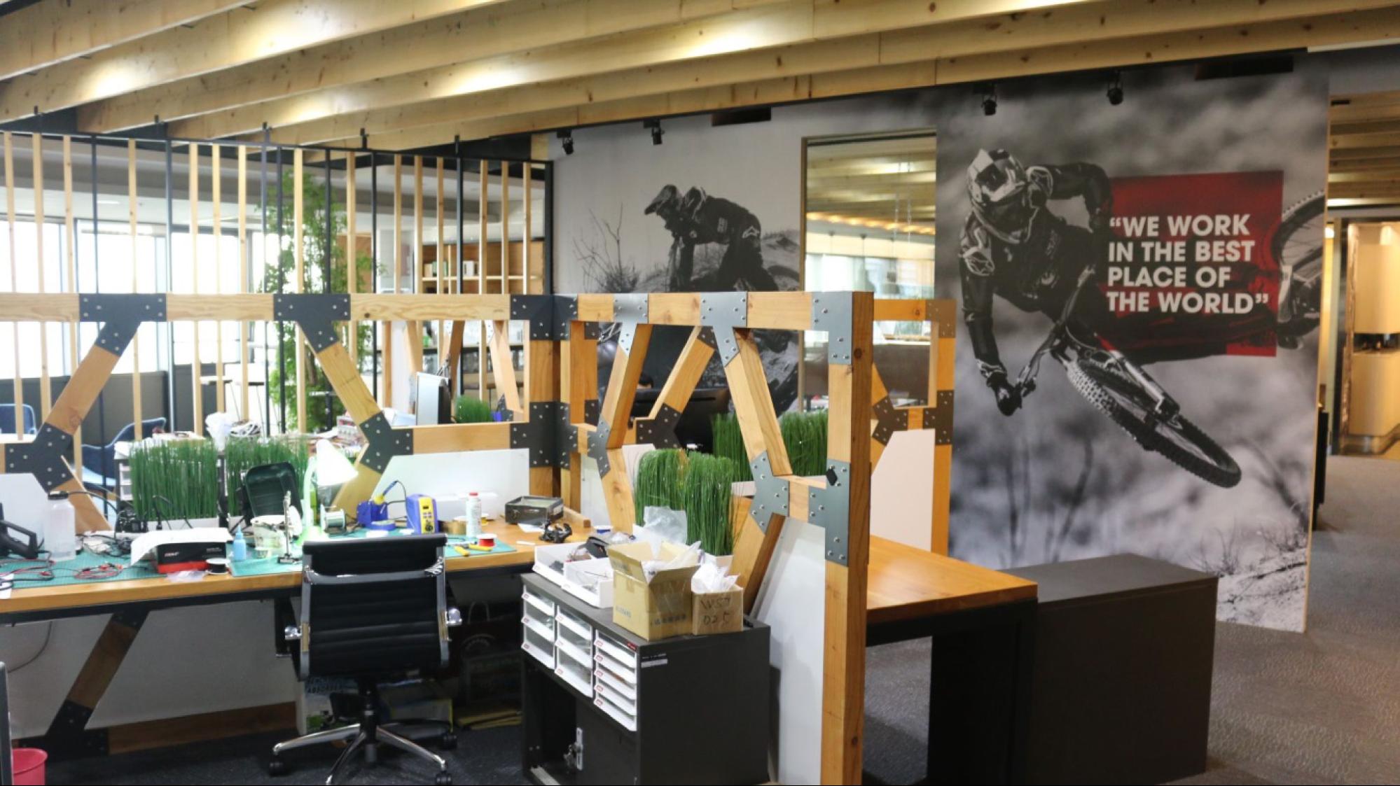 An office interior with wooden-framed partitions, desks, chairs, and large wall murals featuring mountain biking photos and a quote.