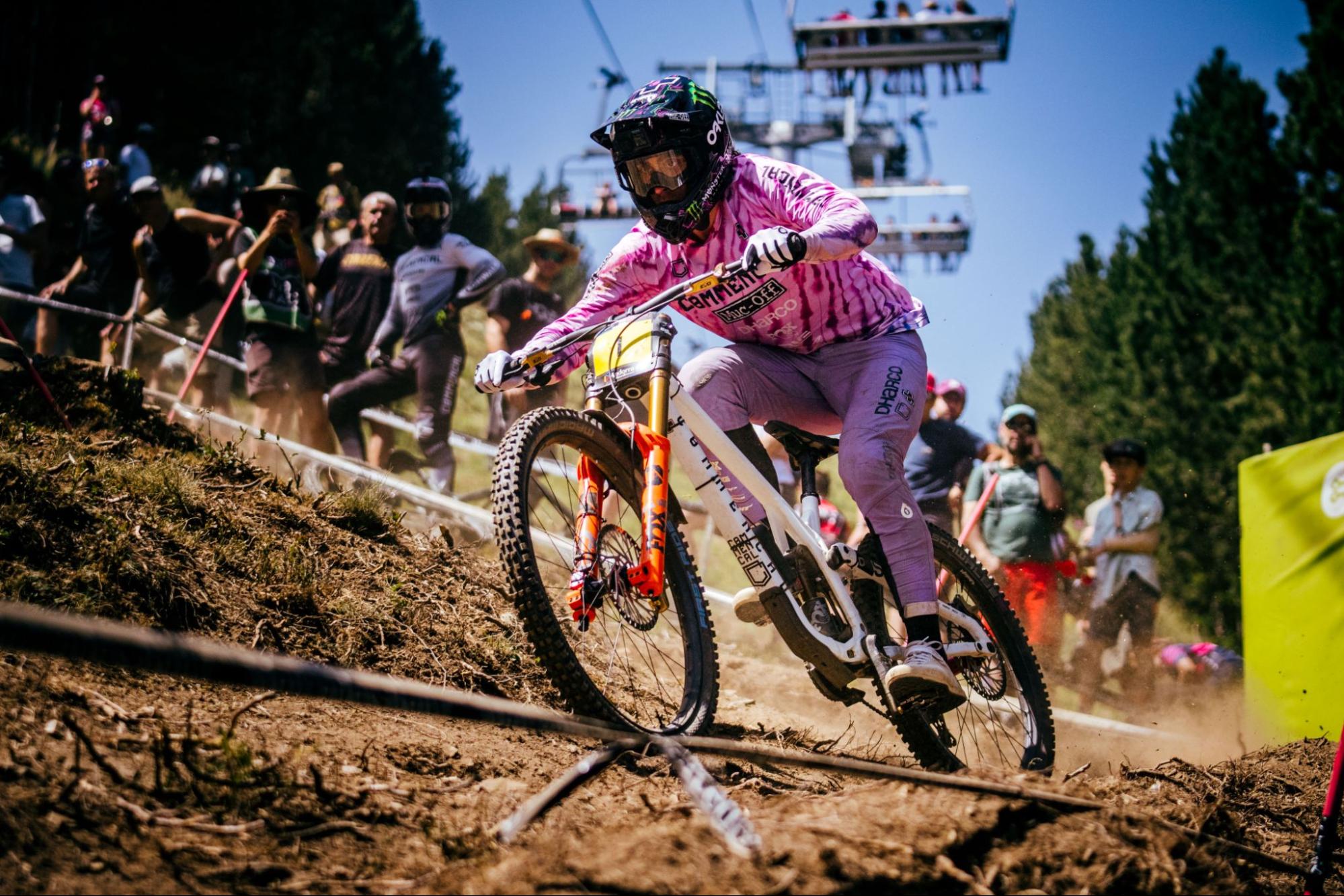 A mountain biker in a pink patterned jersey and purple pants navigates a dirt track at high speed, with spectators and a ski resort lift visible in the background.