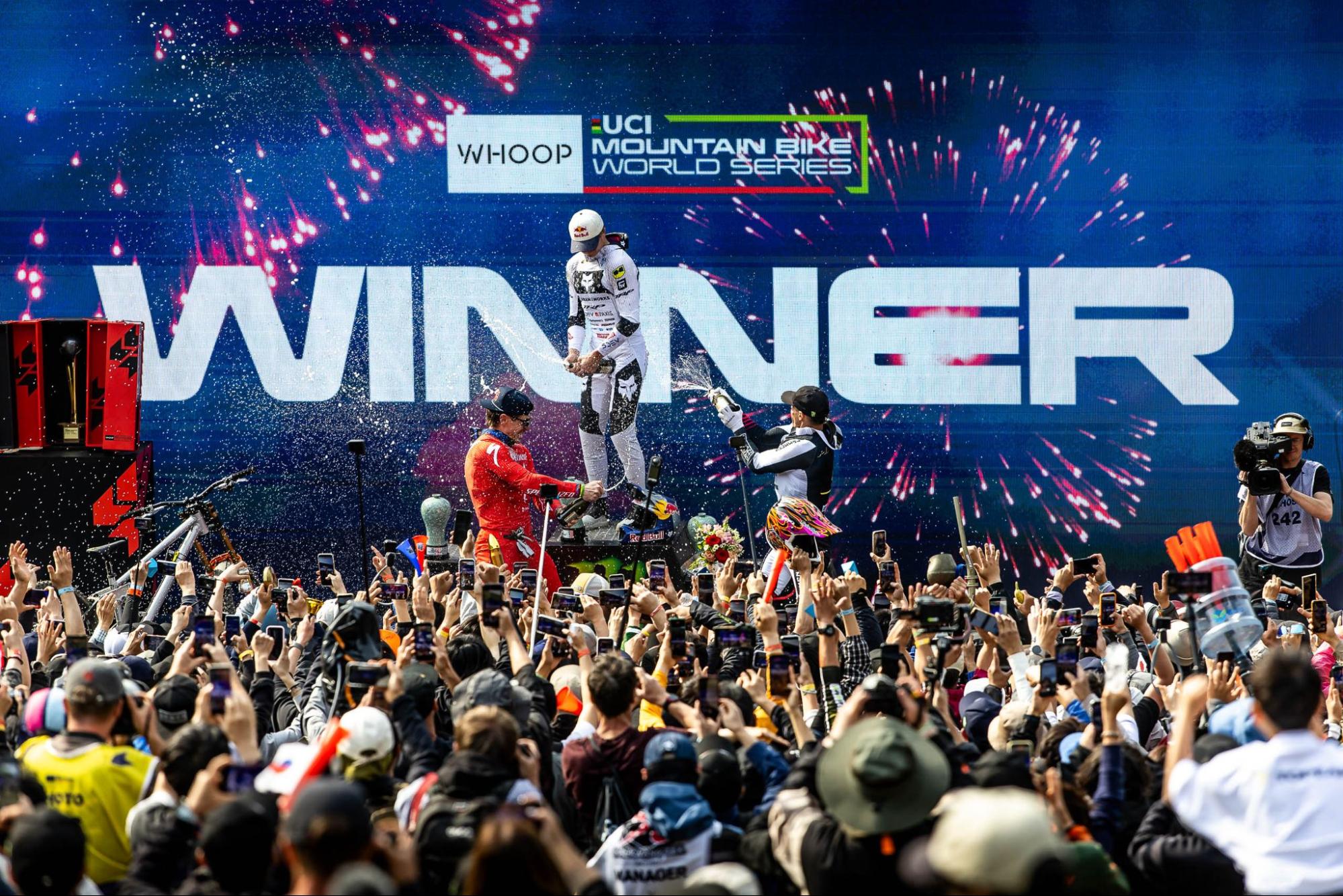 A crowd of spectators cheers on three UCI Mountain Bike World Series riders on a podium during a live event. A large blue backdrop features the text 'WHOOP' and 'WINNER' with fireworks exploding in the background.