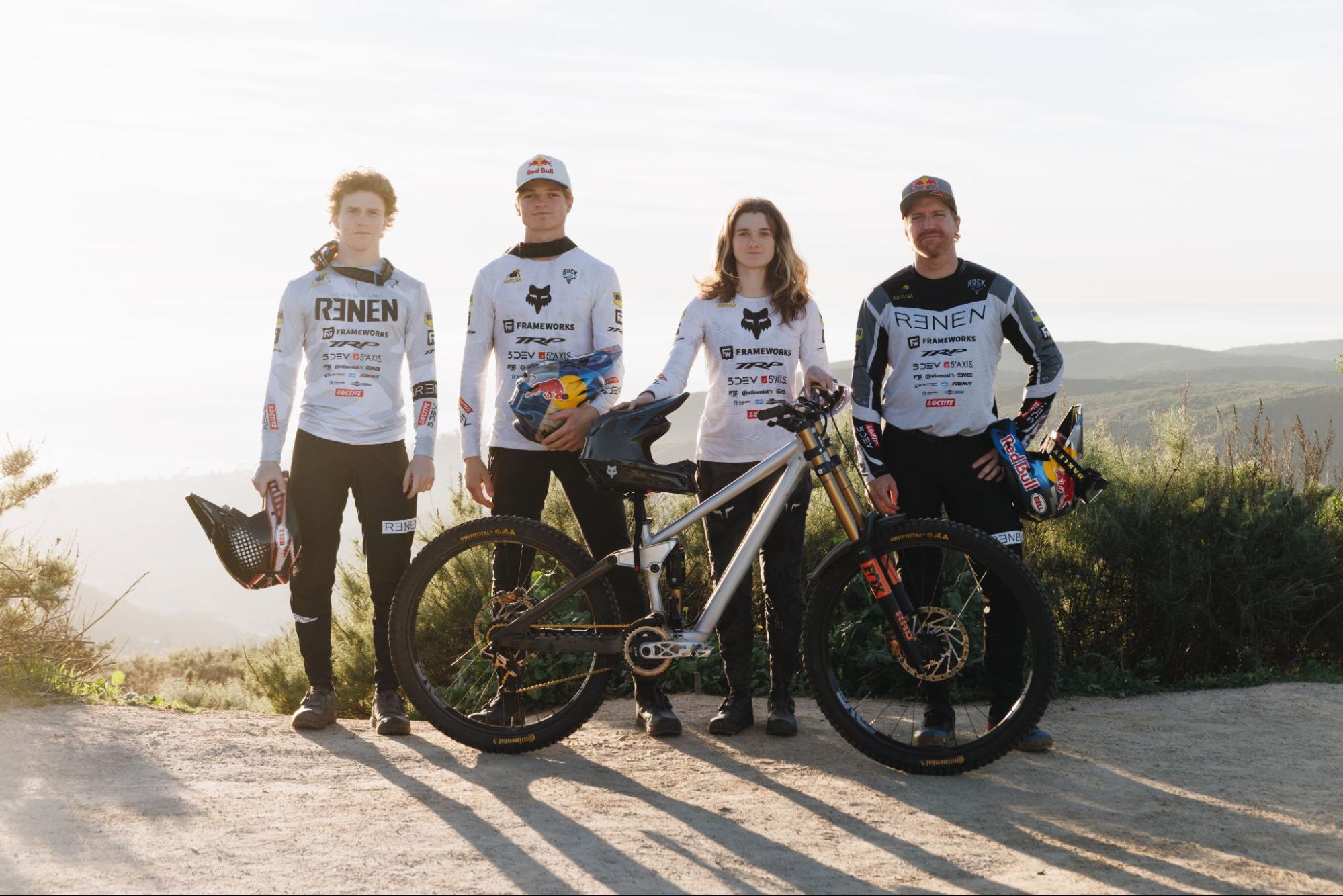 Four mountain bikers standing outdoors on a sunlit dirt path with two full-suspension bikes in front of them. They are wearing matching white and black Reinen cycling jerseys with sponsor logos, helmets, and gloves, set against a blurred natural landscape background.