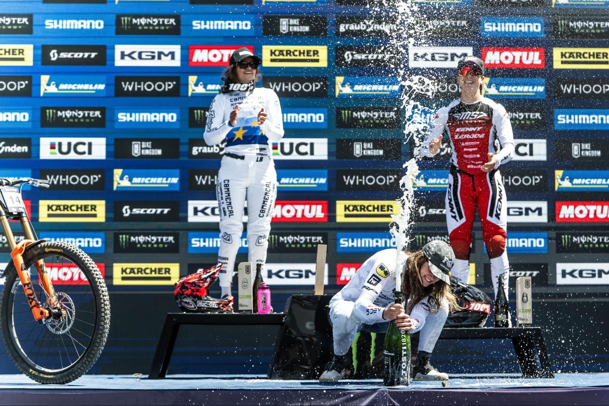 Three mountain bike riders on a podium with champagne spraying, set against a blue sponsor backdrop with logos like Shimano, KGM, and Monster Energy.