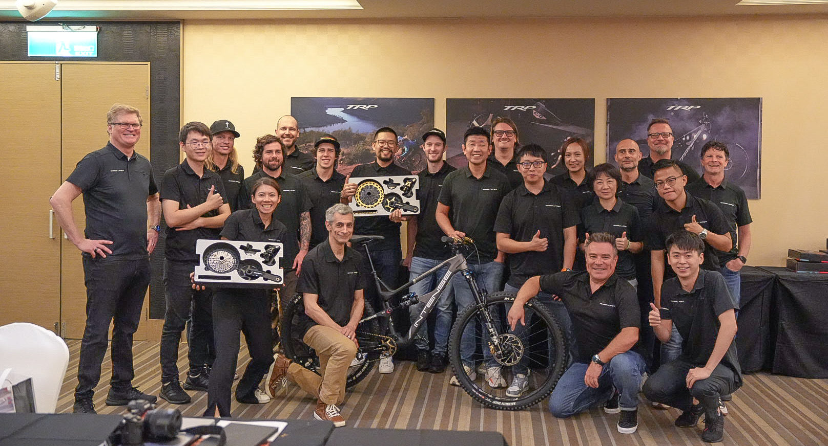 A group of TRP amployees posing together in an indoor office or meeting room, wearing matching black polo shirts. Several individuals are holding product boxes, and a bicycle is visible in the center.