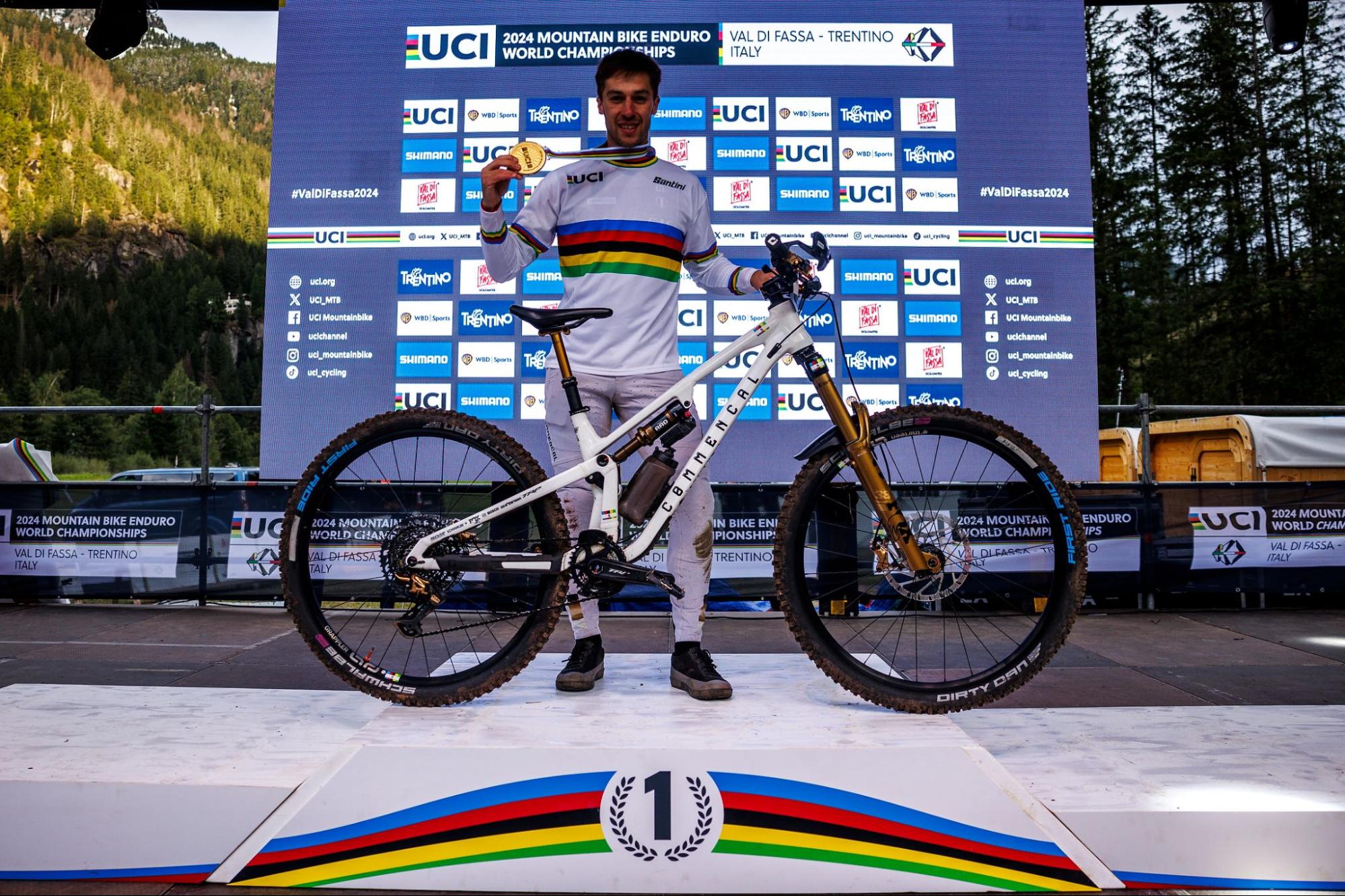 A mountain enduro rider stands on a podium holding a rainbow-colored medal, with a white and gold UCI Enduro Pro mountain bike in front of a blue UCI event backdrop. The rider wears a white jersey with a rainbow stripe and stands on a white platform with a rainbow stripe at the bottom.