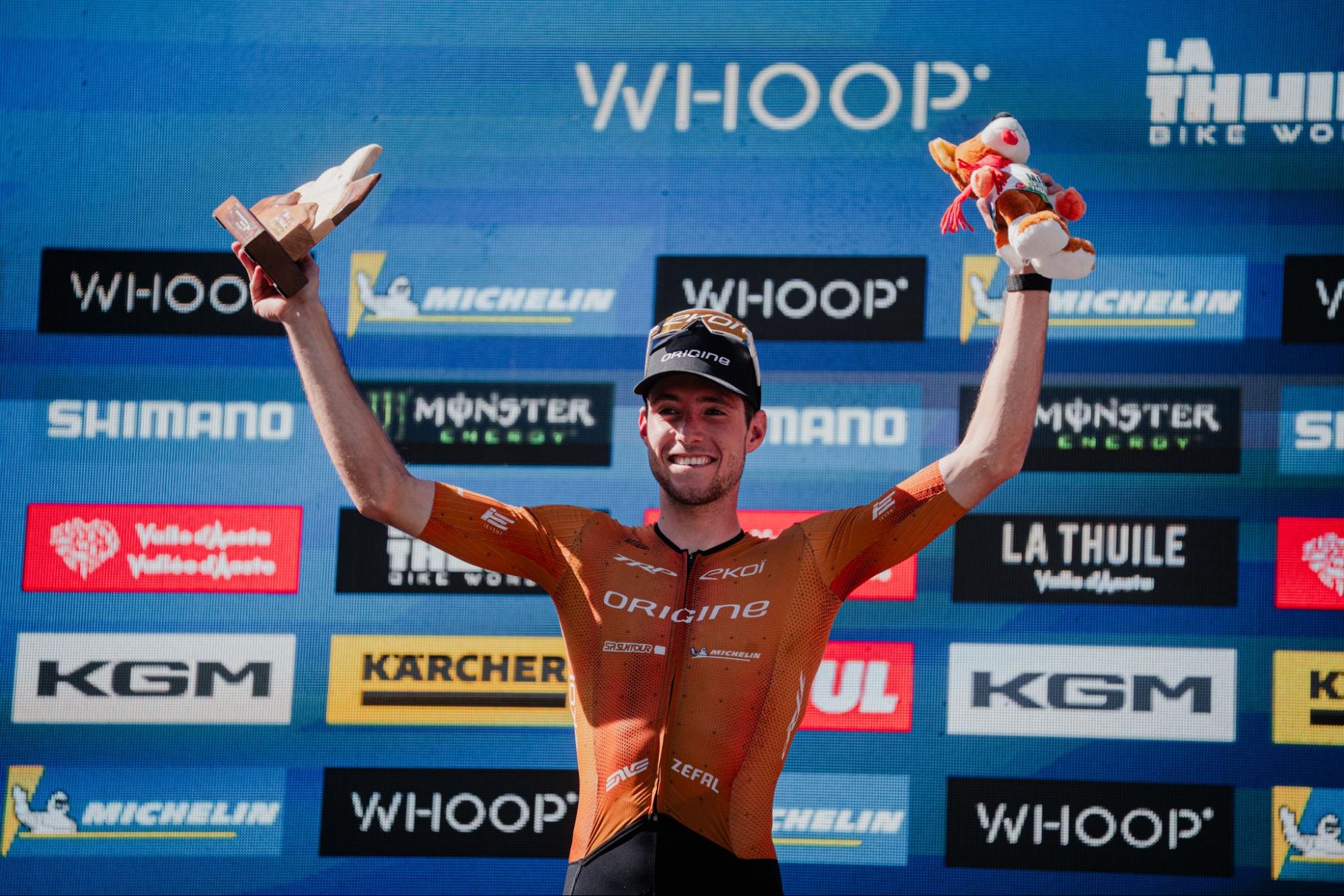A cyclist in an orange and black jersey celebrates on a podium with both arms raised, holding a trophy and a small plush toy in front of a blue sponsor backdrop featuring Whoop, Michelin, Shimano, and Monster Energy.