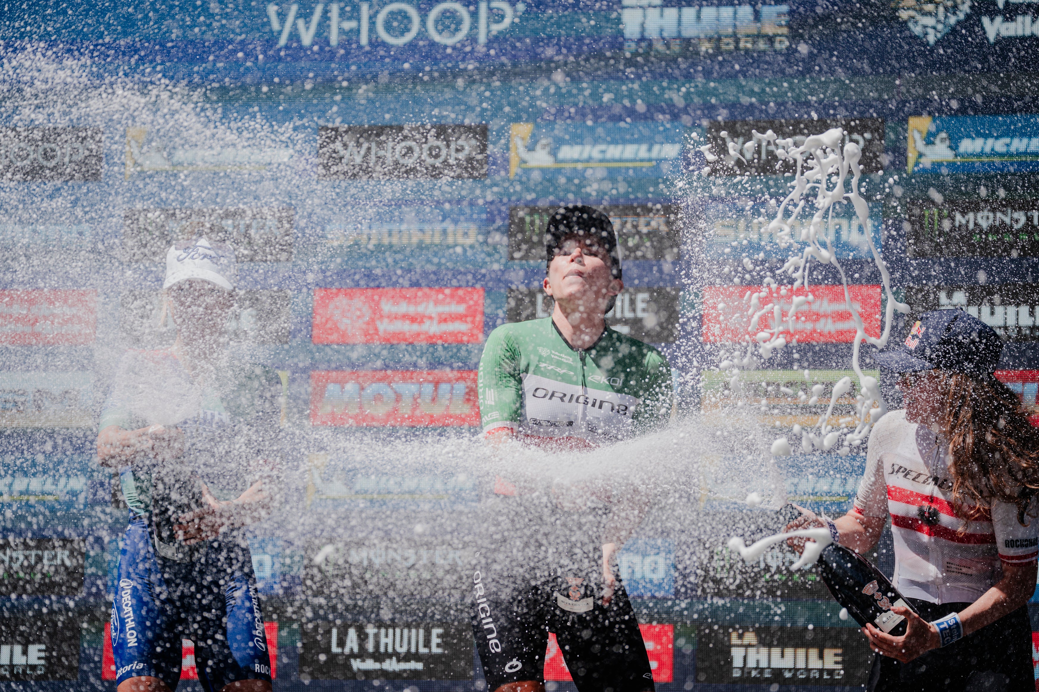 A cyclist in a green jersey celebrates on a podium with champagne spraying across the scene, with other riders and branded sponsor banners in the background.