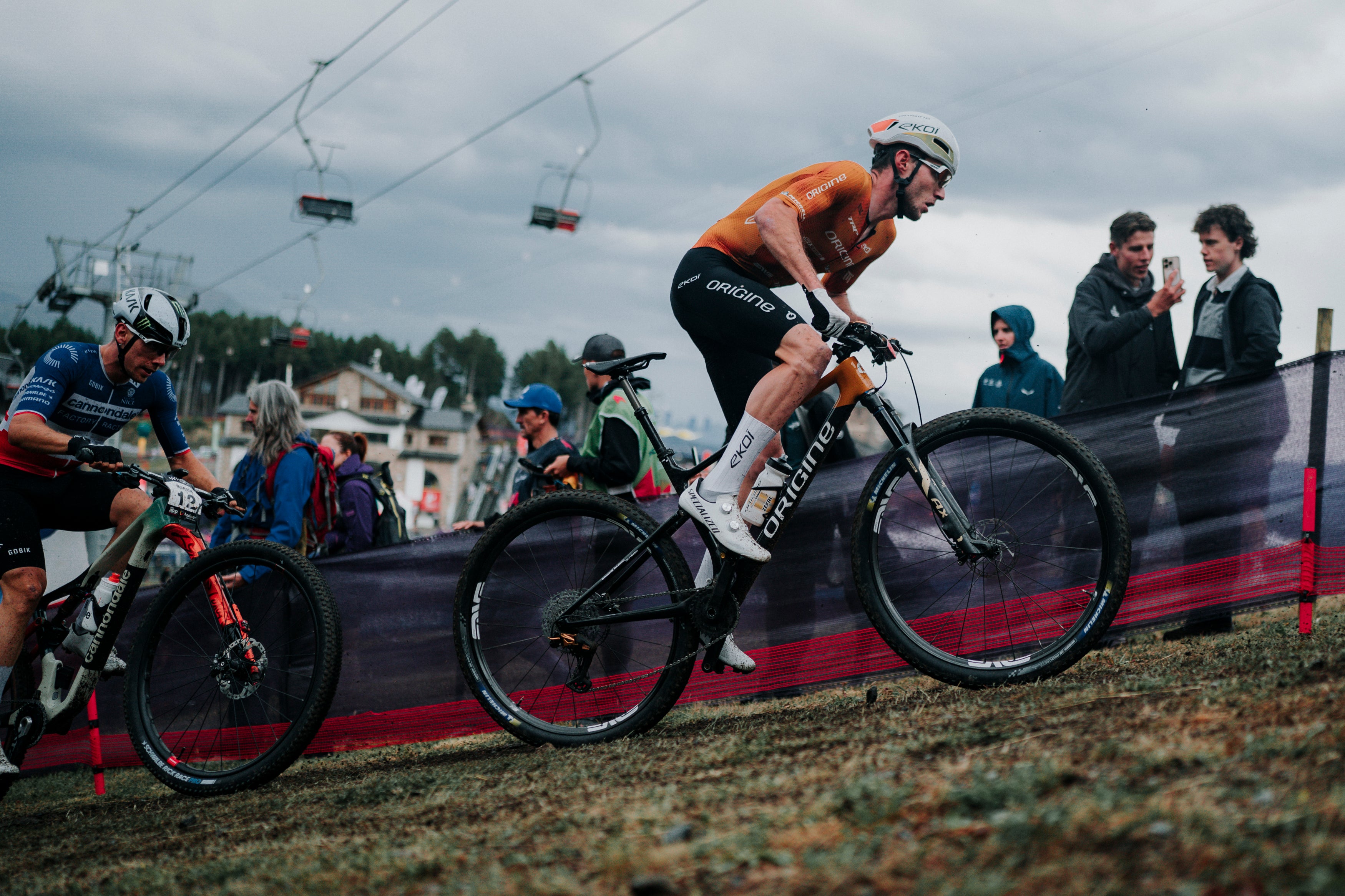 A mountain biker in an orange jersey and black shorts rides a black and silver Santa Cruz full-suspension bike during an outdoor race, with other racers, spectators, and ski lifts visible in the background under an overcast sky.