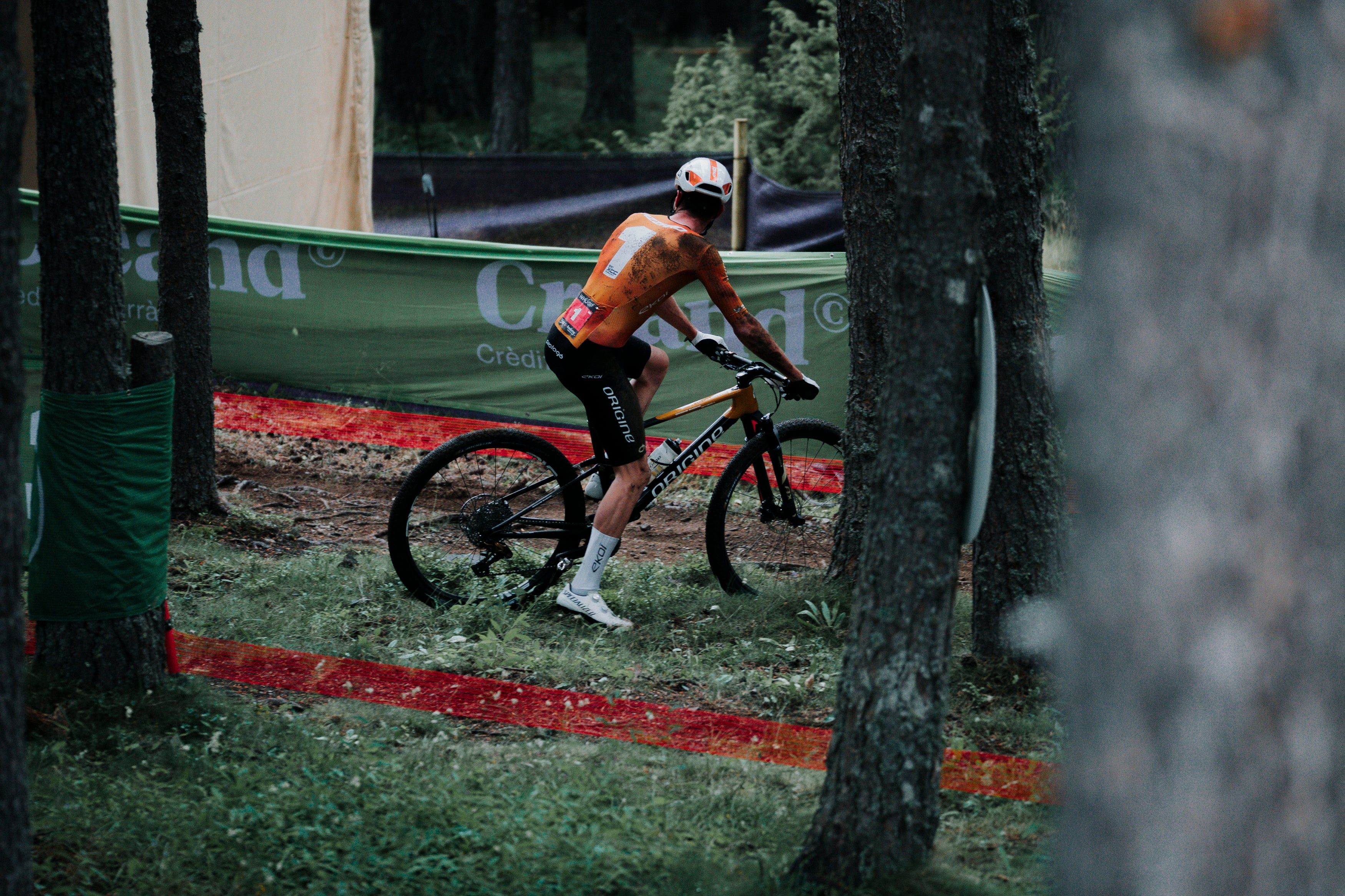 A mountain biker wearing an orange and white jersey, black shorts, and white socks rides a black bike through a wooded course with a red boundary line and trees in the background.