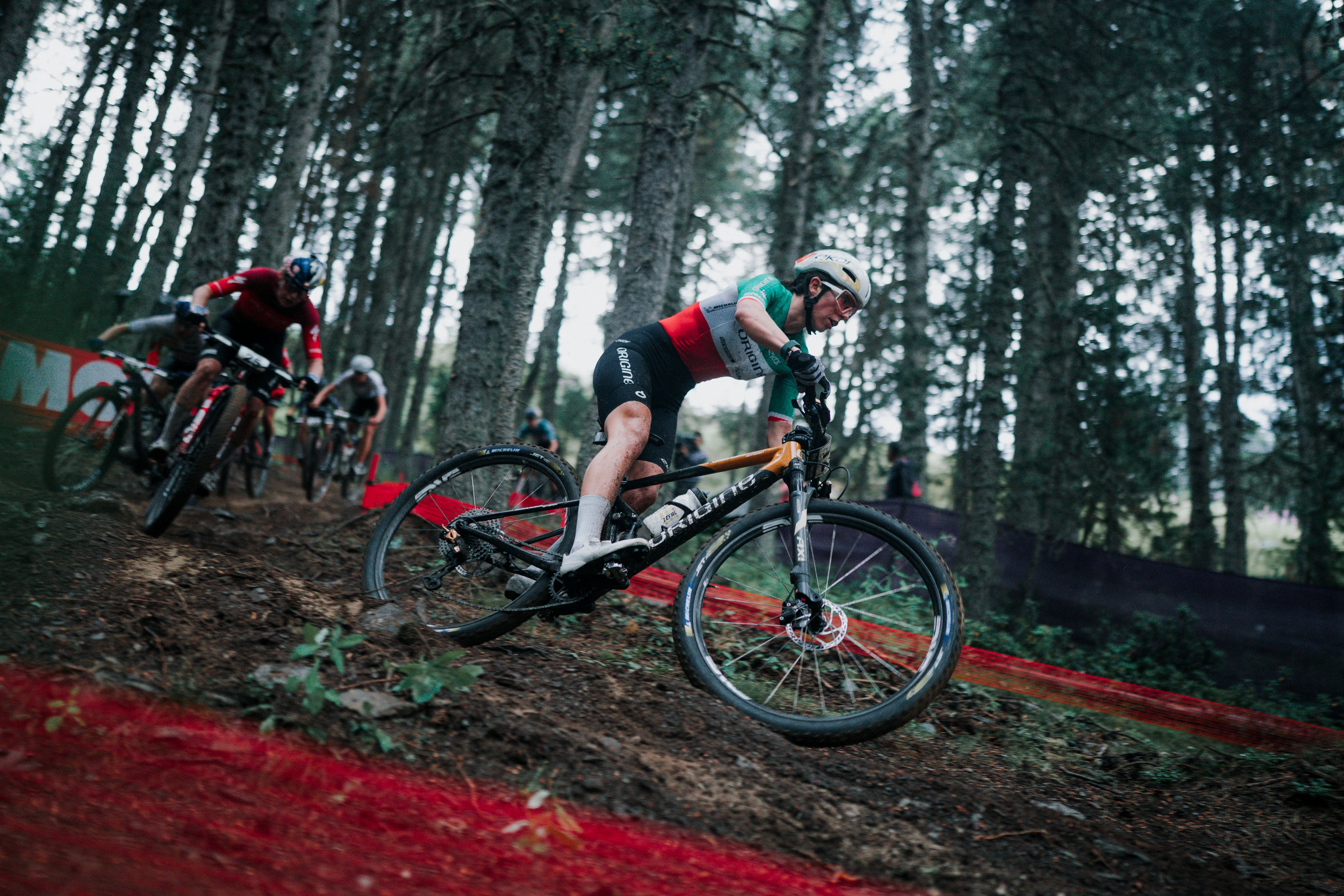 A mountain biker in a forest course wearing a red, black, and green jersey, riding a black and orange Santa Cruz mountain bike on a dirt track with other racers in the background.