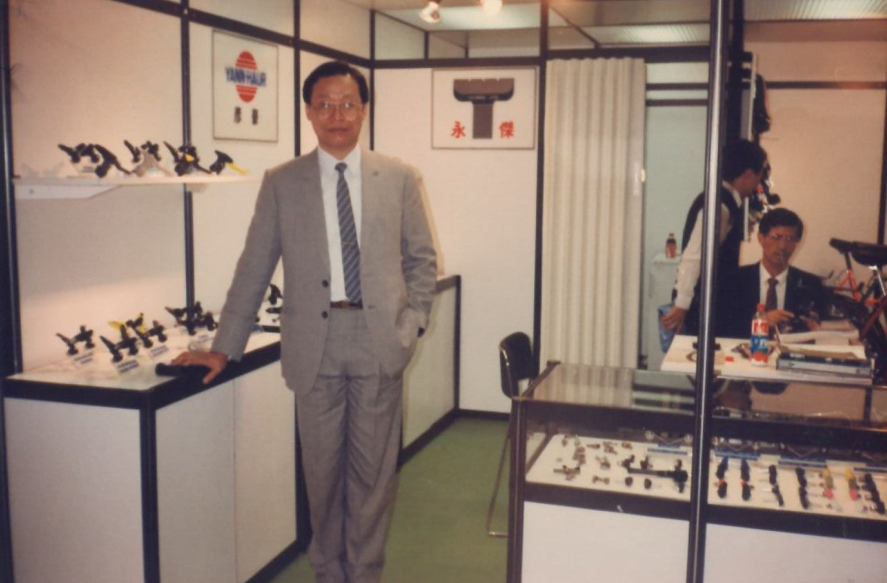 A man in a light gray suit and striped tie stands in a glass-walled exhibition booth, with display cases containing small products behind him and another man seated at a desk to the right.